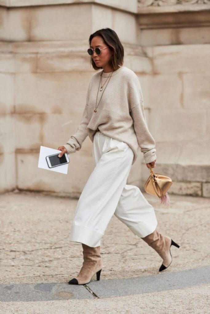 Mulher veste pantacourt branca com trico bege, óculos de sol, bota cano longo e finaliza com a bolsa clutch doura
